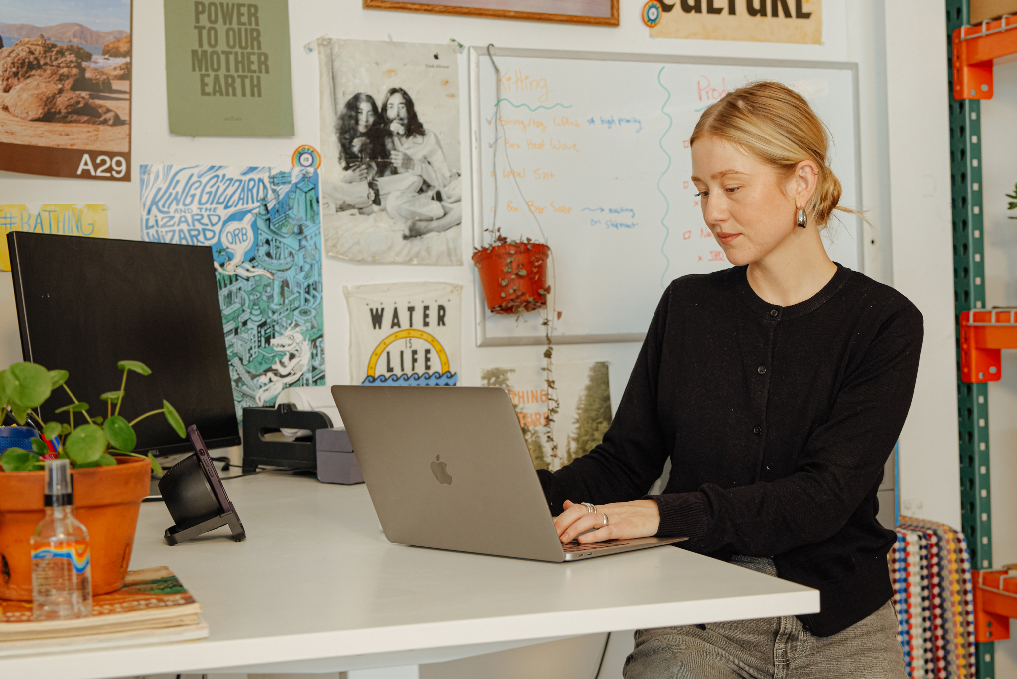 a blonde woman sits at a desk, looking at a laptop