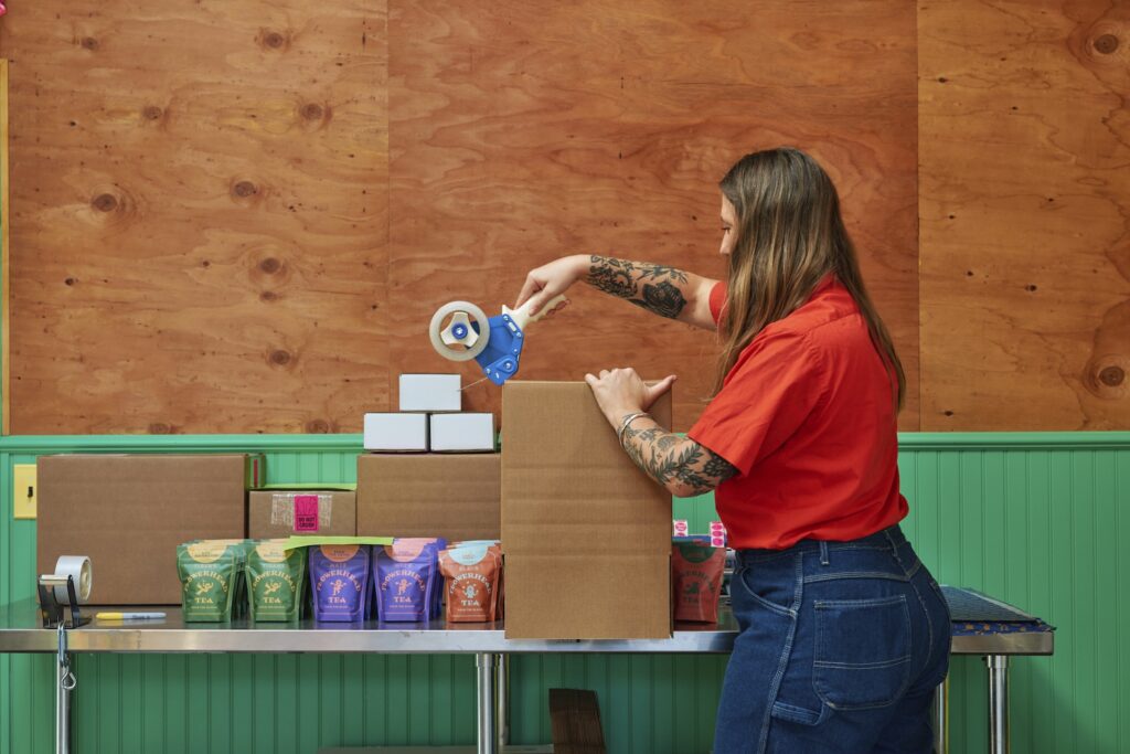 Eine Frau im roten Hemd packt Beutelchen in einen großen Pappkarton ein. Später kann sie die durchshnittliche Bestellwert oder AOV von diesen Produkten kalkulieren.