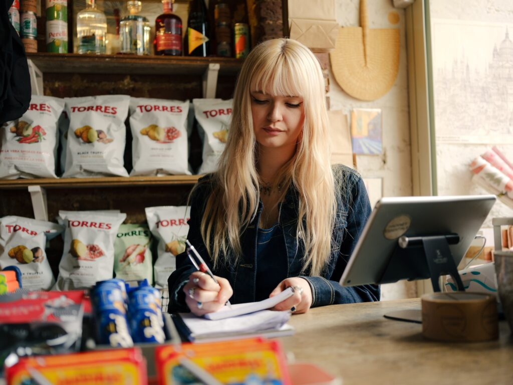 Eine blonde Frau macht eine manuelle Rechnung für Kleinunternehmer, mit Chipsverpankungen im Hintergrund.